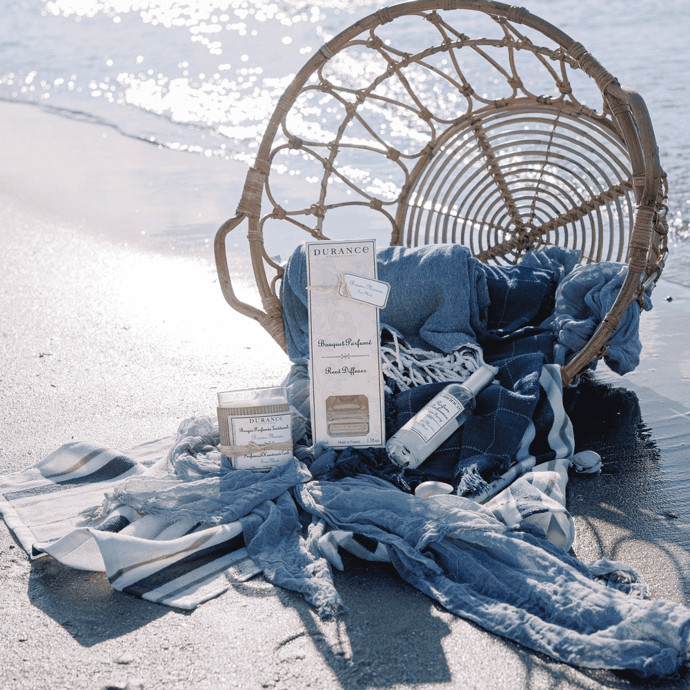 bougie artisanale brume marine sur la plage avec la mer autour et d'autres parfums Durance ainsi qu'un panier avec du linge bleu 